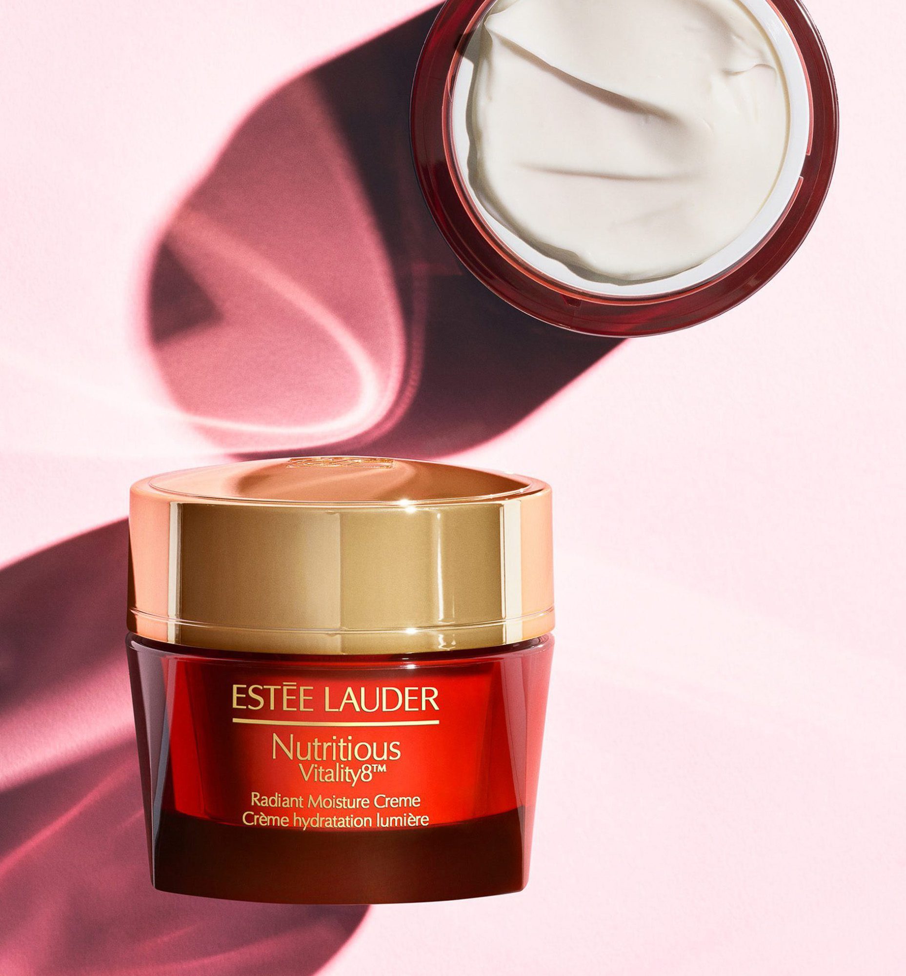 Red and gold Nutritious Vitality8 moisture creme jar beside an open jar of white cream on pink, for Estée Lauder.