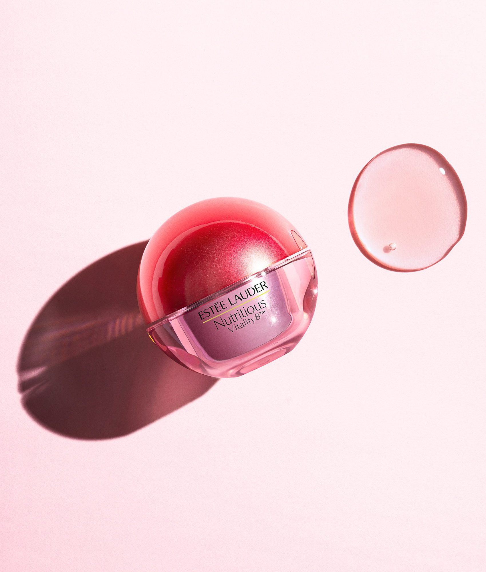 Still life of a red spherical Nutritious Vitality8 jar beside a clear gel swatch on a pale pink surface, for Estée Lauder.
