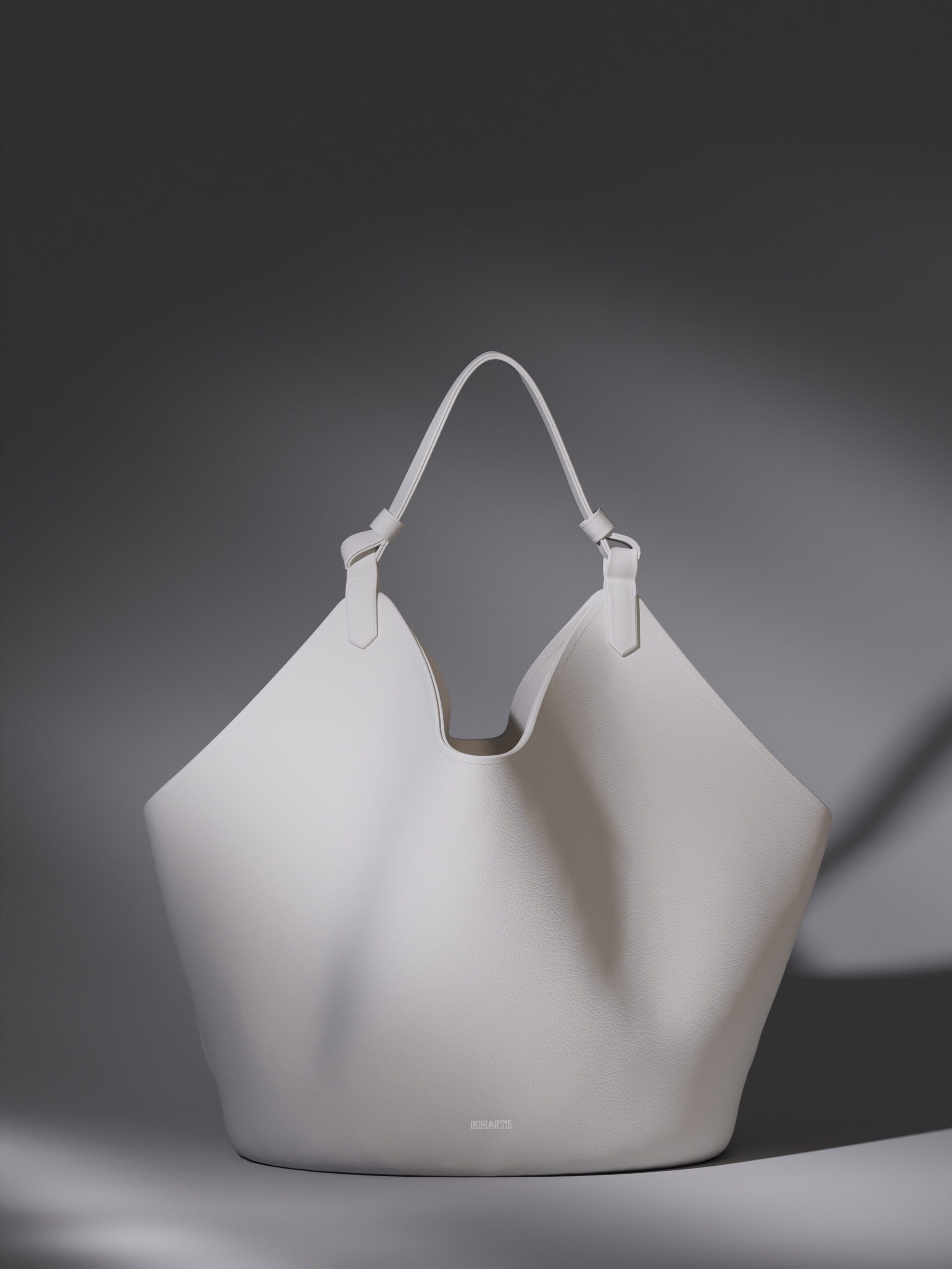 White leather tote bag with knotted shoulder strap under soft shadowed light on a grey backdrop, for Khaite.