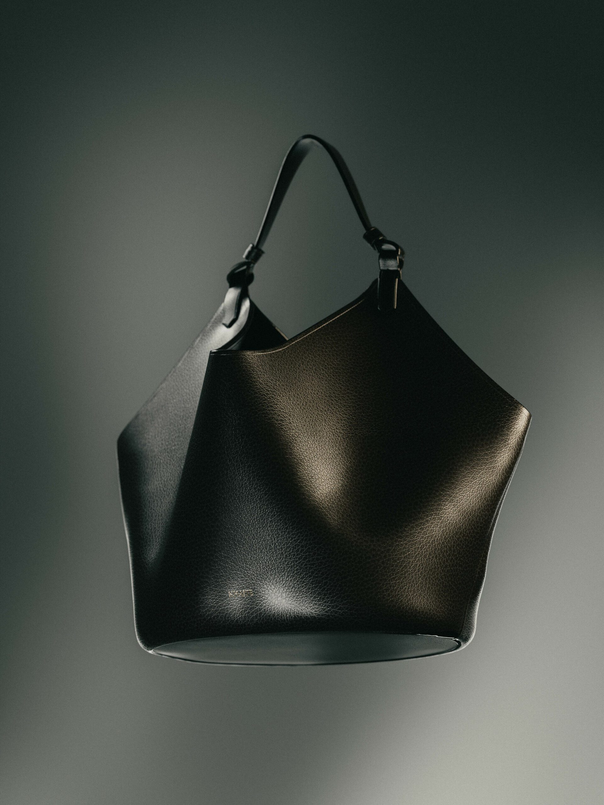Black grained leather bucket bag with a single top handle, lit softly against a grey backdrop, for Khaite.