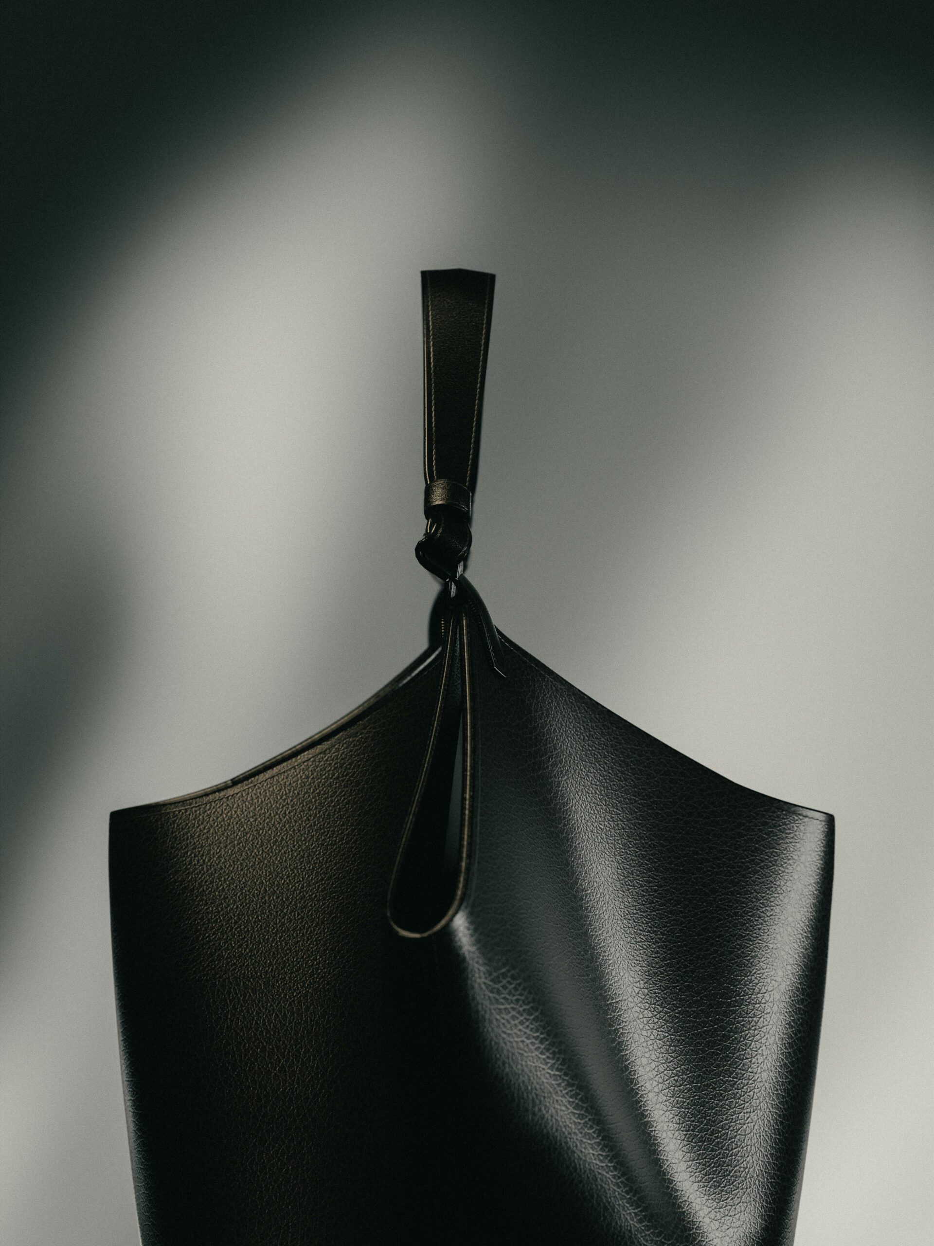 Black grained leather tote with a knotted strap, lit against a grey wall with soft shadow, for Khaite.