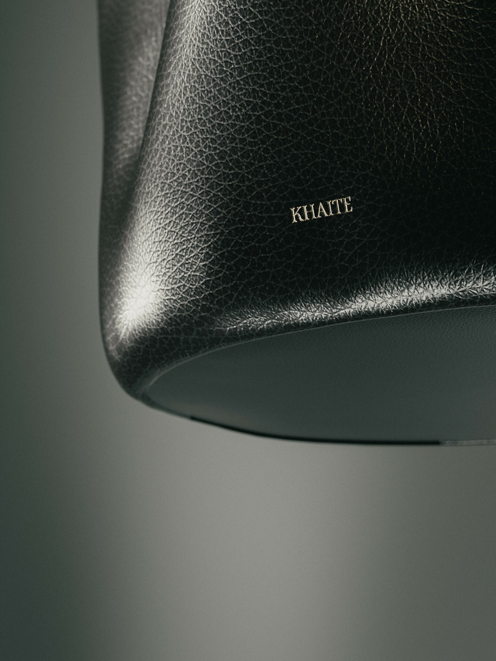 Close-up of a black pebbled leather bag with a gold-stamped logo against a soft grey backdrop, for Khaite.