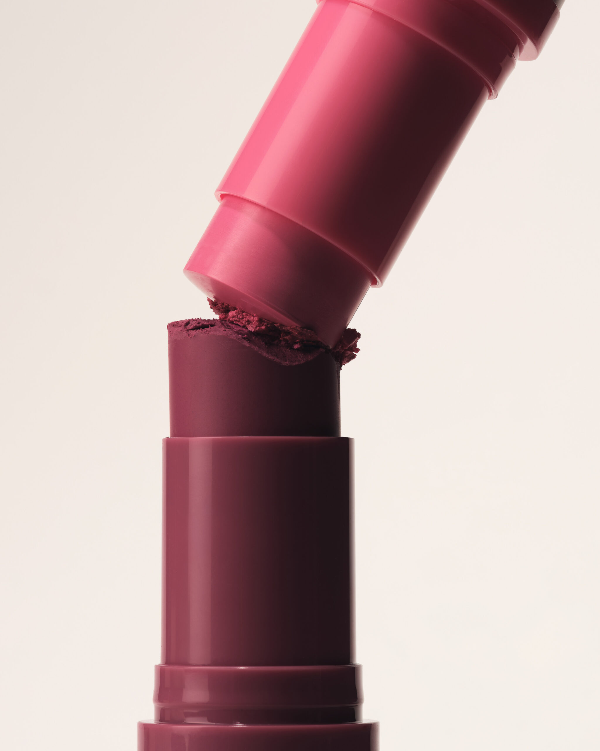 Still life of a pink lipstick tube pressing into a crumbling plum lipstick bullet on a cream background, for Kylie Cosmetics.