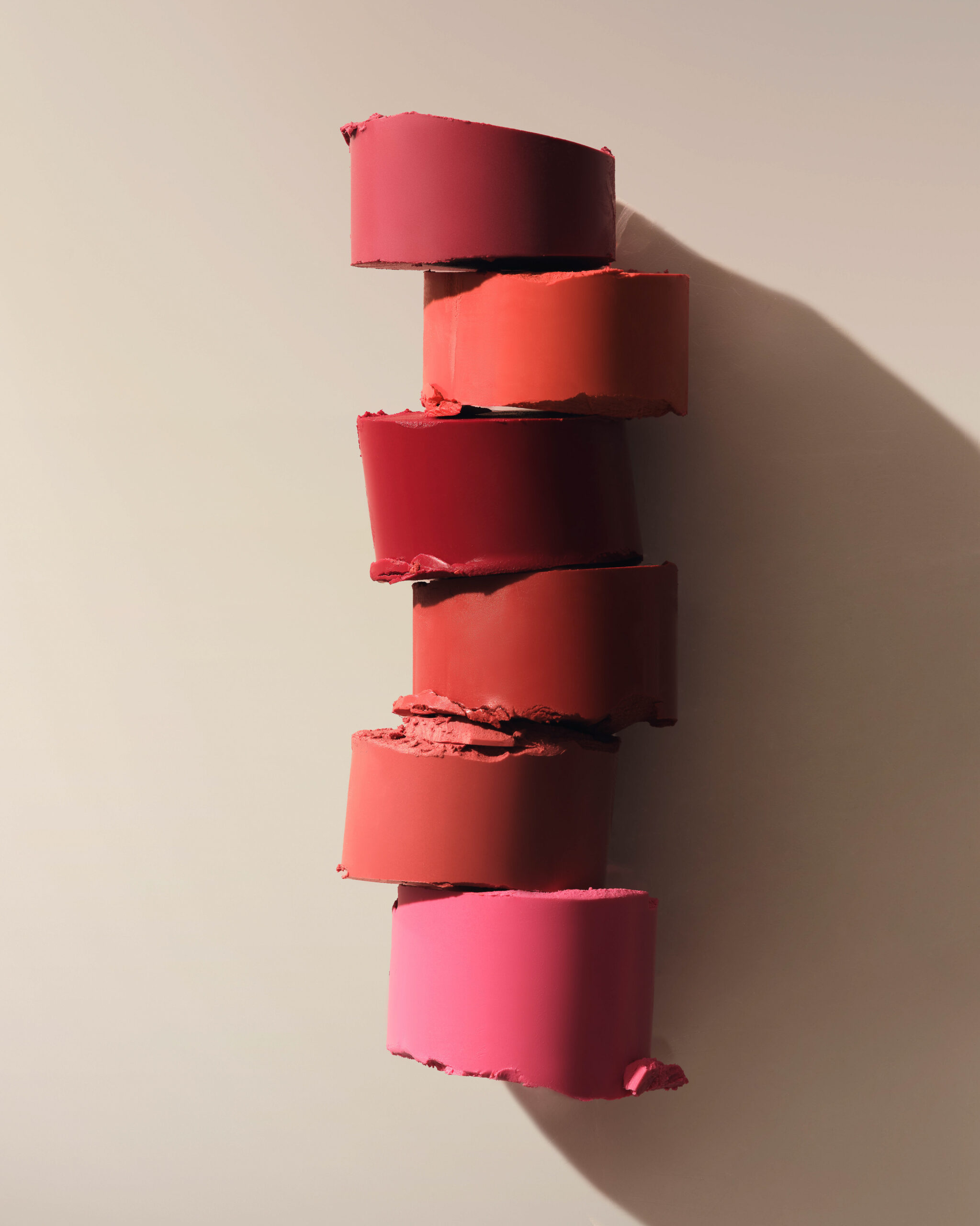 Still life of six lipstick slices in pink, red and coral shades stacked on a beige wall with a long shadow, for Kylie Cosmetics.