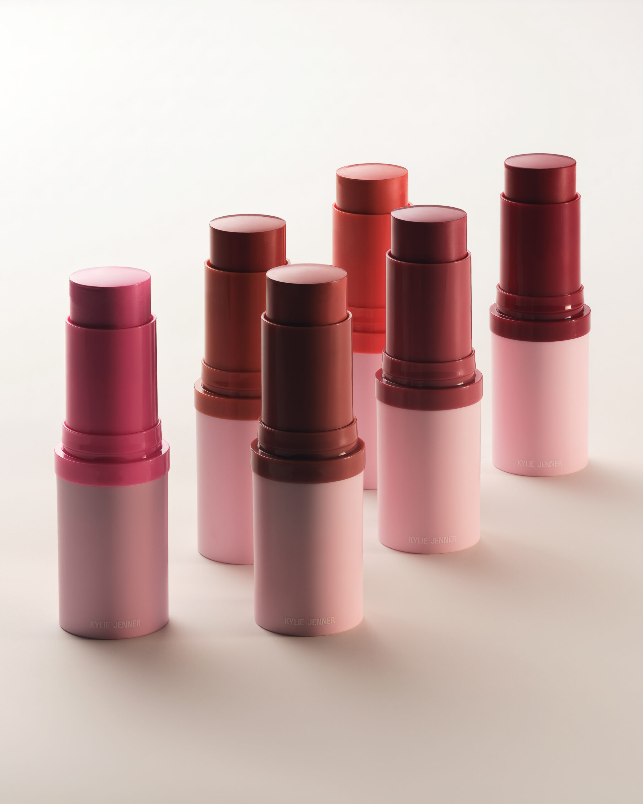 Six cream blush sticks in pink, berry and brick red shades with pale pink bases on a cream backdrop, for Kylie Cosmetics.