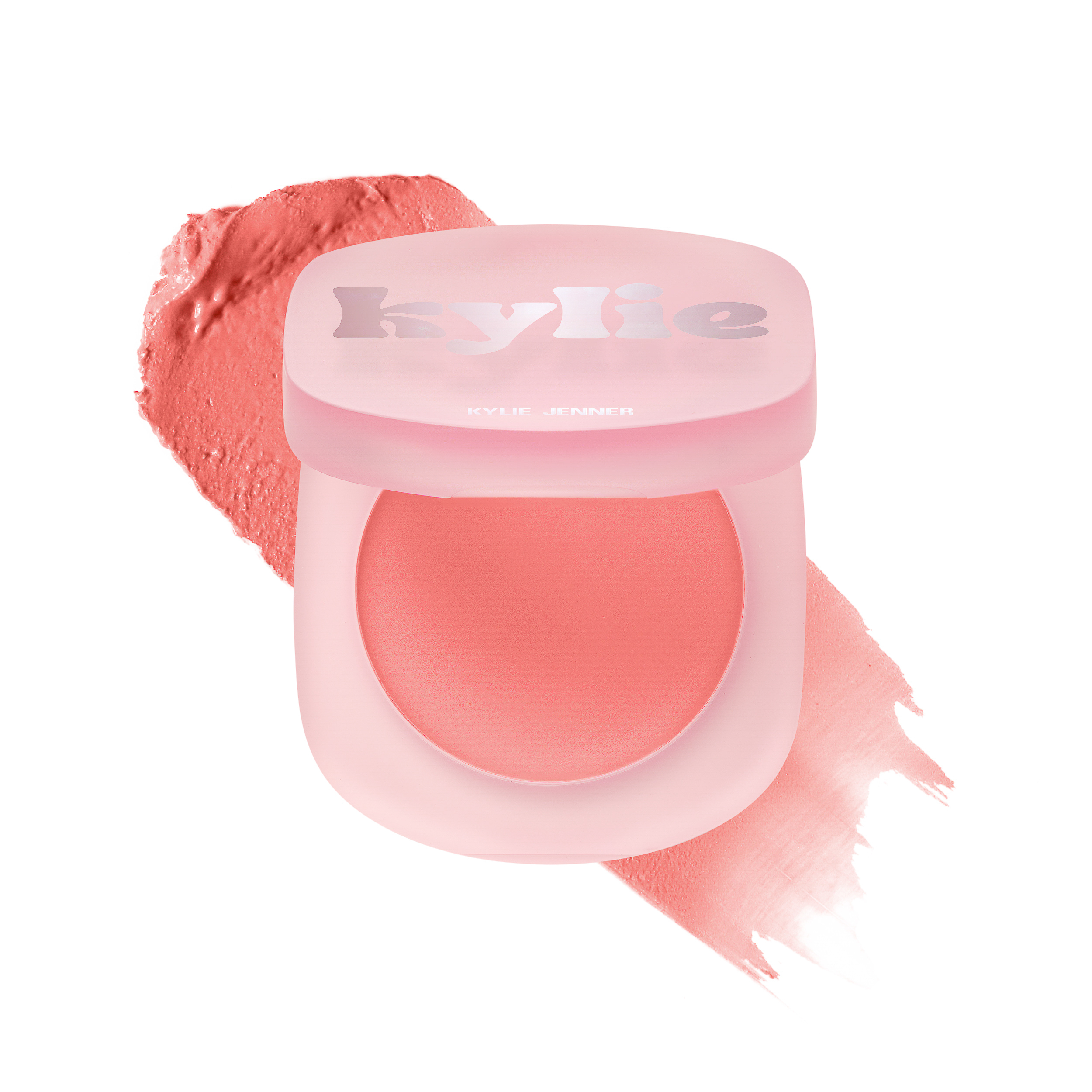 Open frosted pink cream blush compact with a coral swatch smeared behind it on white, a beauty still life, for Kylie Cosmetics.