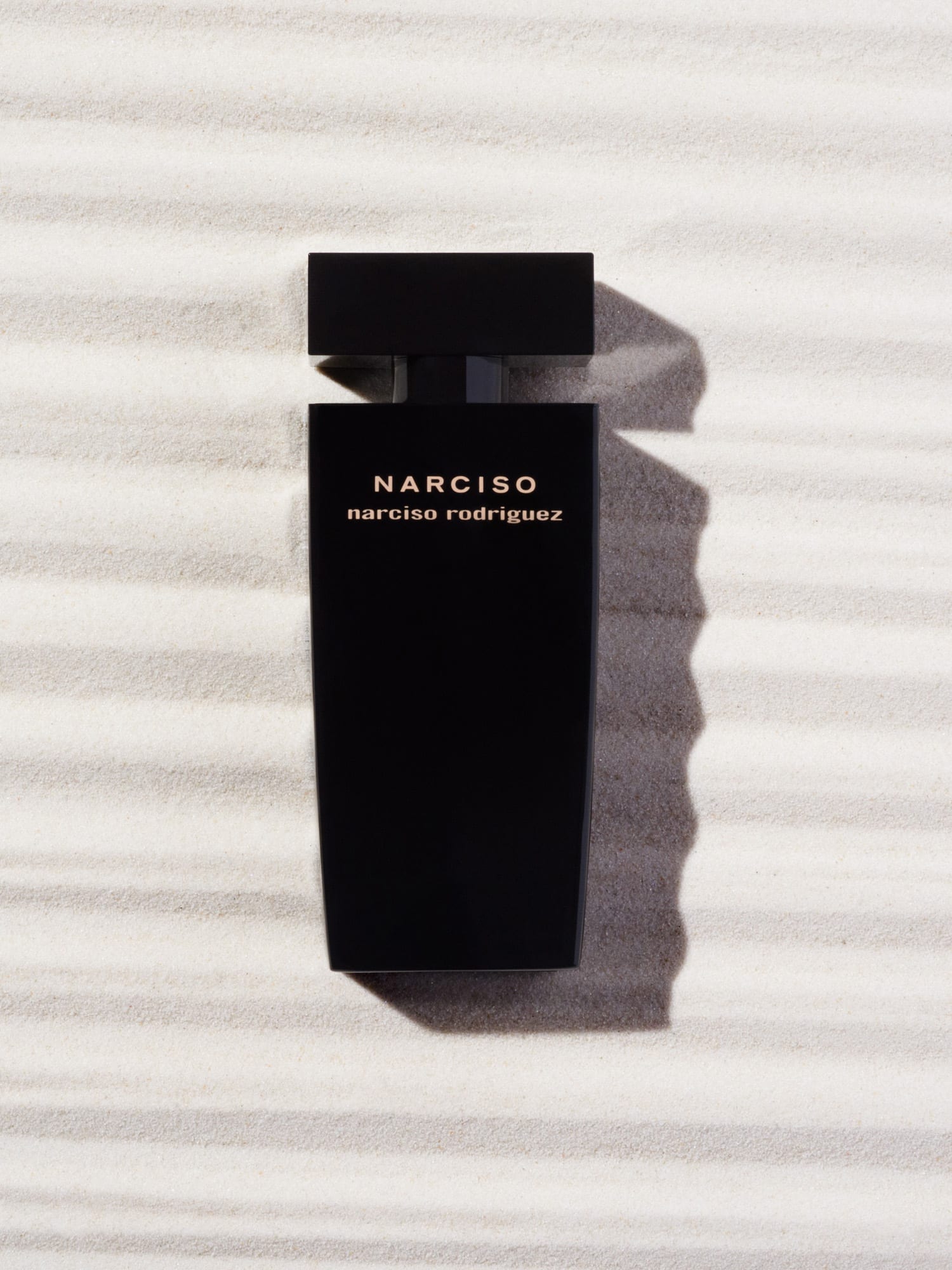 Black NARCISO fragrance bottle lying on rippled white sand under hard sunlight with a long shadow, for Narciso Rodriguez.